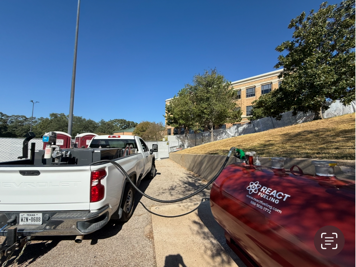 Two parked pickup trucks on a sunny dirt path with hoses/cables stretched between them near a hillside building