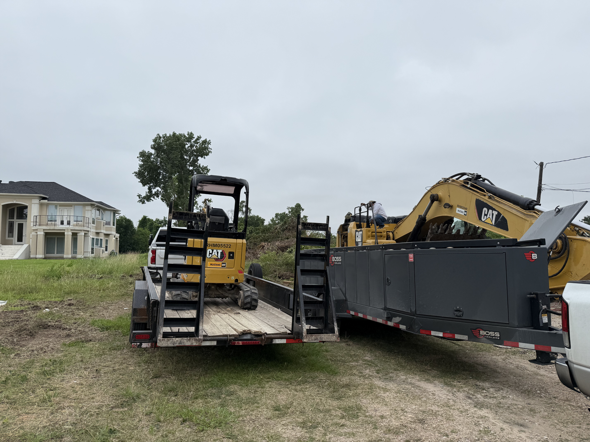 Construction equipment on trailers in a residential lot, with a yellow excavator and a house in the background