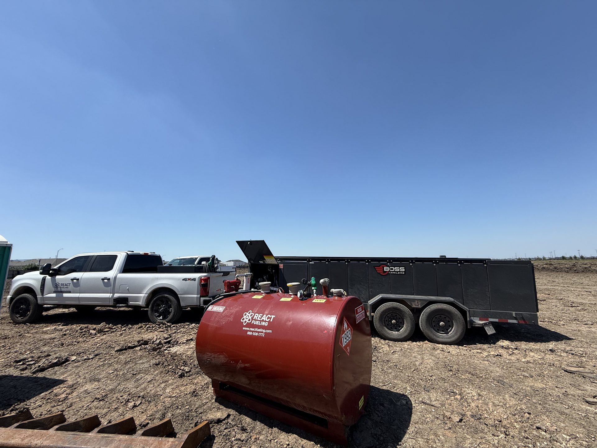 White pickup and black dump trailer on gravel beside a red industrial tank under a clear blue sky
