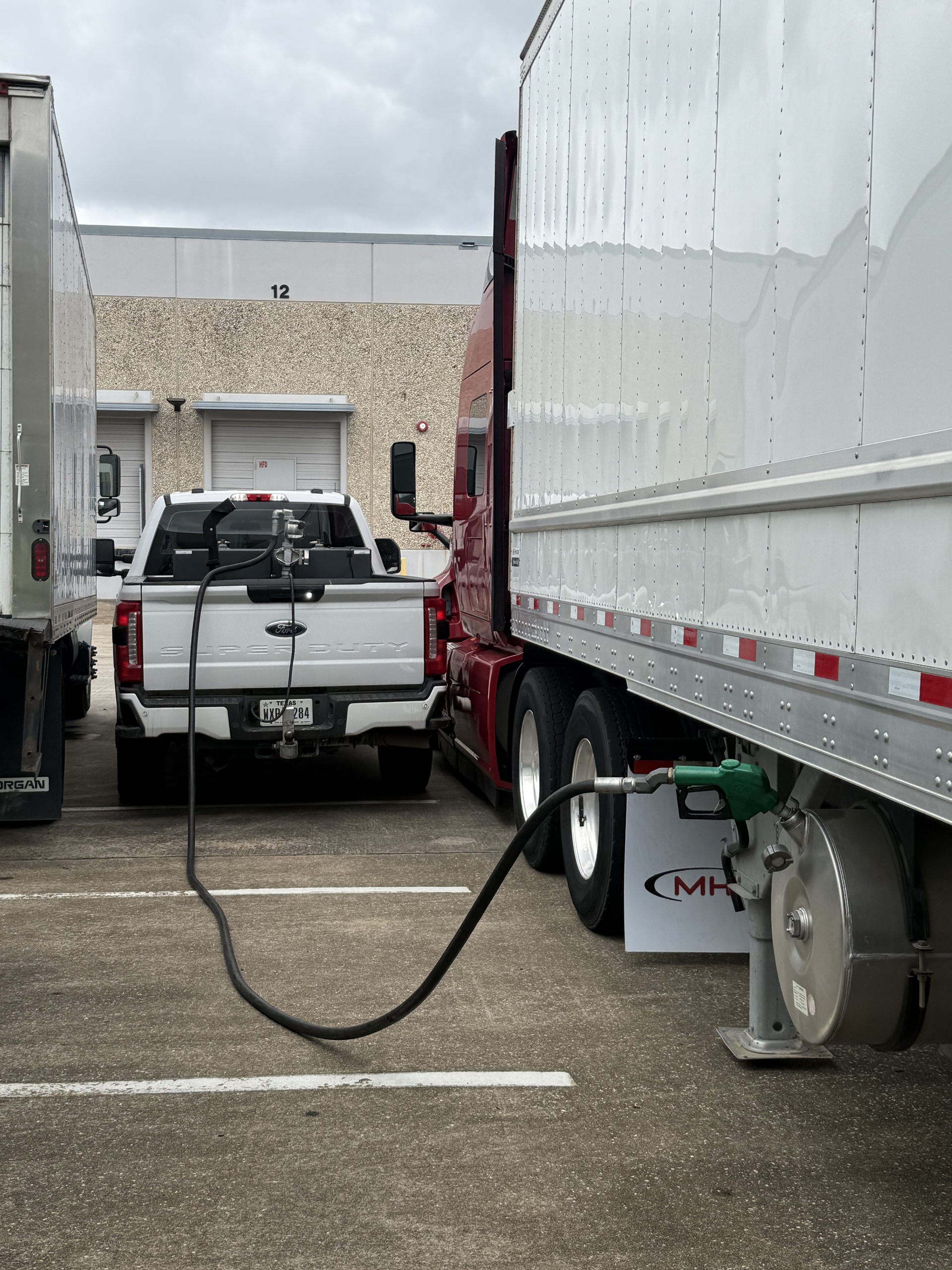 White box truck fueling a white pickup truck in a parking lot, with a fuel hose between them.