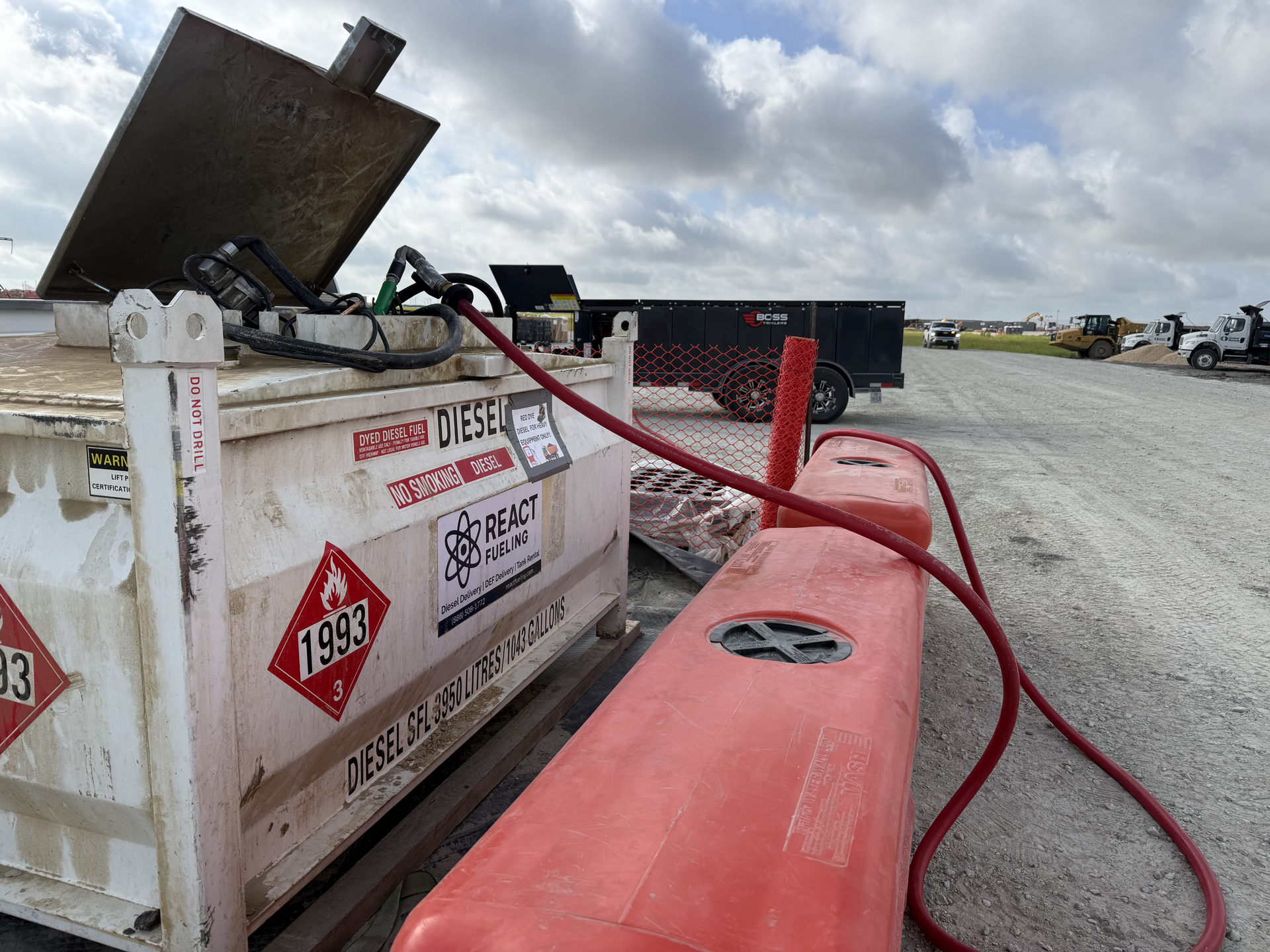 Red fuel tank in gravel lot beside a white dumpster and black equipment under a cloudy sky