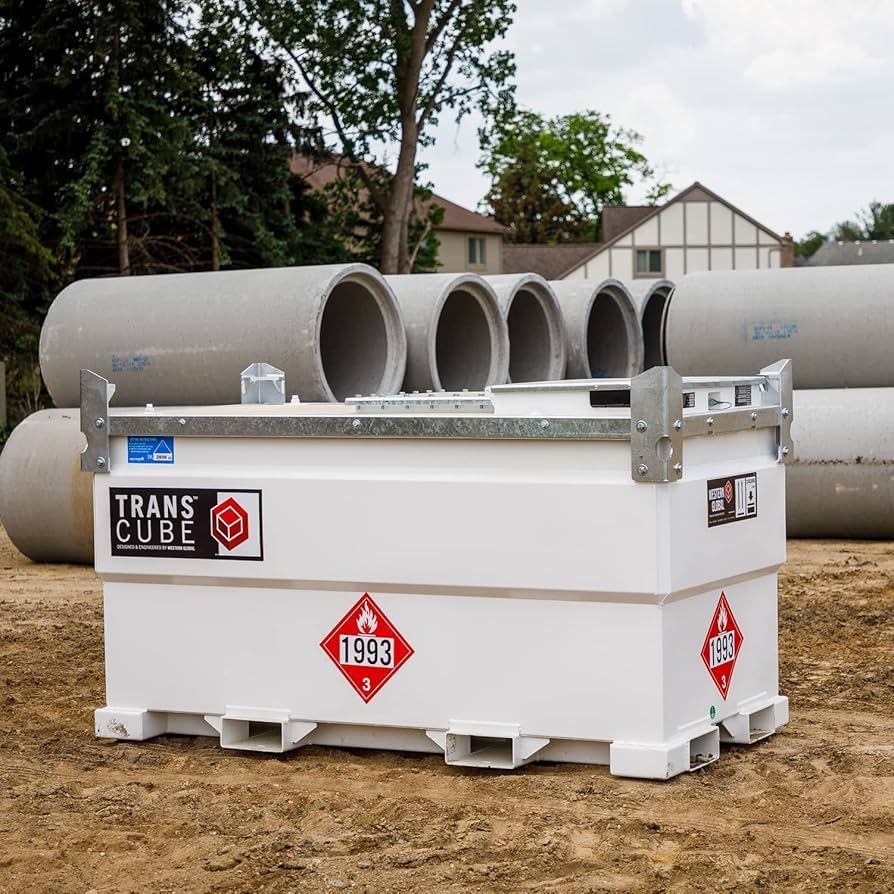 White TransCube storage tank with hazard signs, set outdoors on a dirt lot near large concrete pipes.
