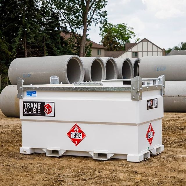 White TransCube storage tank with hazard signs, set outdoors on a dirt lot near large concrete pipes.