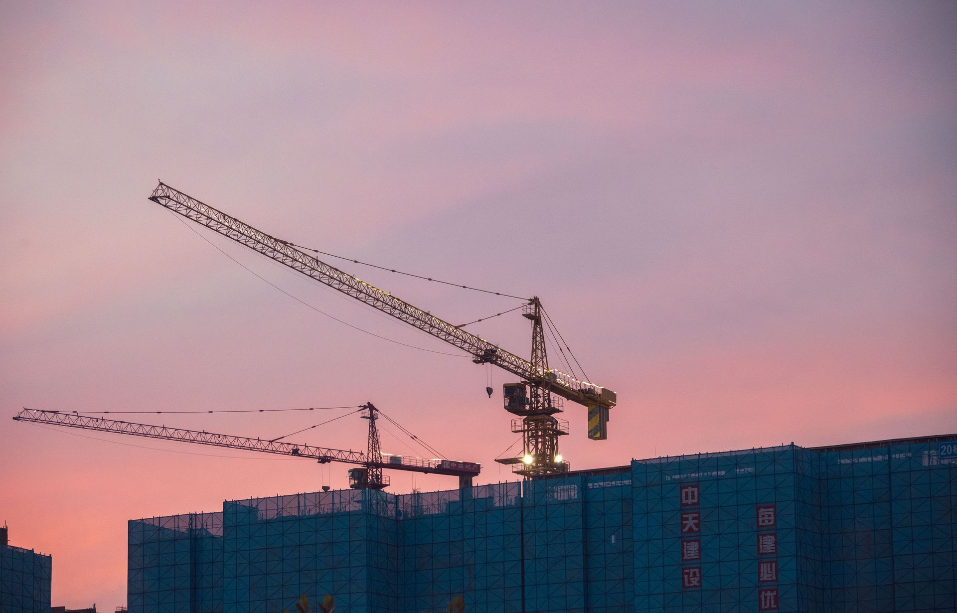 Construction cranes silhouetted against a pink sunset sky over a building site