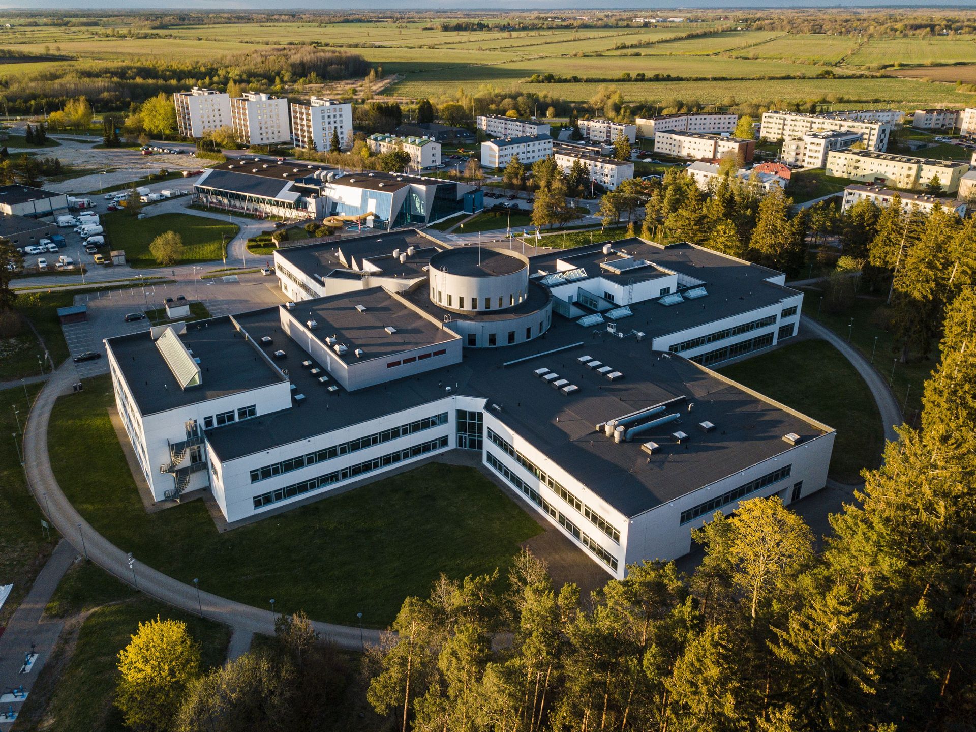 Aerial view of a modern white office complex with a circular central building, surrounded by trees and fields.