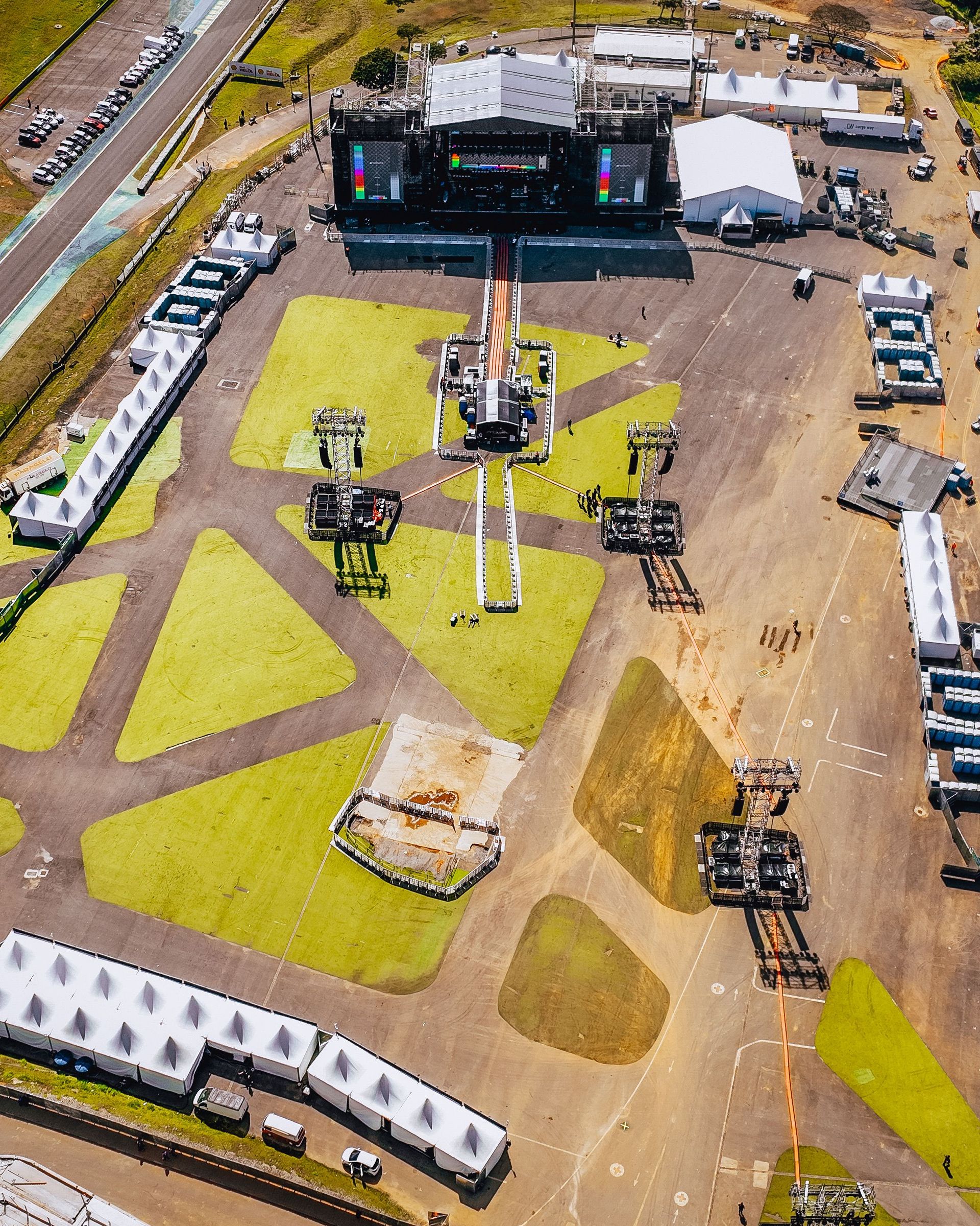 Aerial view of an outdoor event site with a large stage, tents, and vehicles on a grassy field