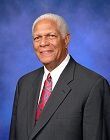 Studio portrait of a man in a dark suit, white shirt, and red tie against a blue background