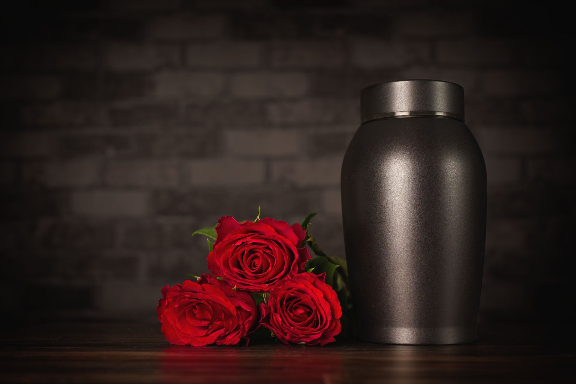 Red roses beside a matte black urn on a dark table against a brick wall.
