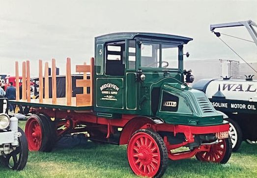A vintage green truck with a red chassis and wooden stake bed sits on a grassy field, next to a white tanker truck.