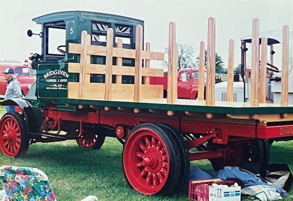 A vintage dark green flatbed truck with red wheels and a natural wood rack, parked on grass at an outdoor show.