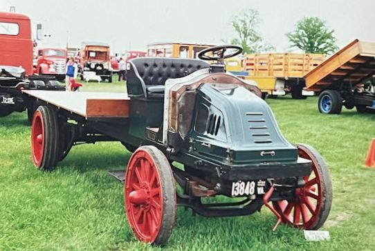 A restored dark green antique flatbed truck with bright red wheels parked on a grassy field at a car show.
