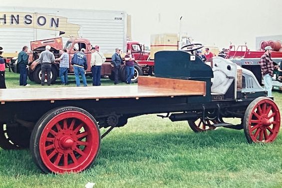 A vintage truck with a flat wooden bed and bright red wheels parked in a grassy field with people standing nearby.