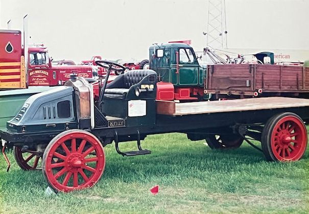 An antique flatbed truck with a black cab, wooden bed, and prominent red spoked wheels, parked on a grassy field.