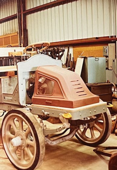 A partially assembled vintage car chassis with spoked wheels, a light blue dashboard, and a terracotta-colored hood in a shed.