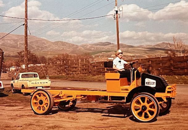 A person in a cowboy hat sits on a restored yellow vintage truck chassis with large spoked wheels in a rural setting.