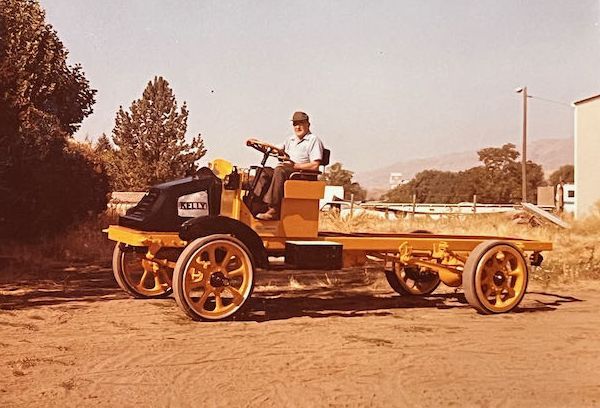 A person sits on a yellow vintage truck chassis with large spoke wheels in a dry, outdoor setting.