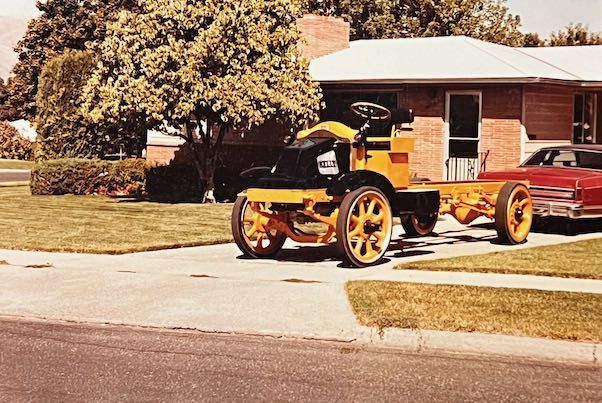A yellow, vintage truck chassis sits in a residential driveway next to a parked red car.