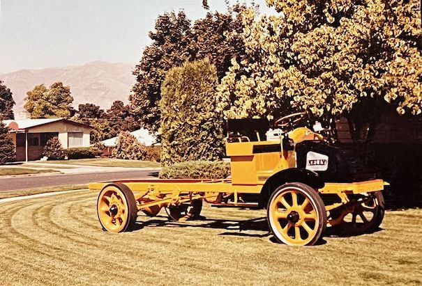 A yellow vintage truck chassis sits on a suburban lawn in front of a house and trees.