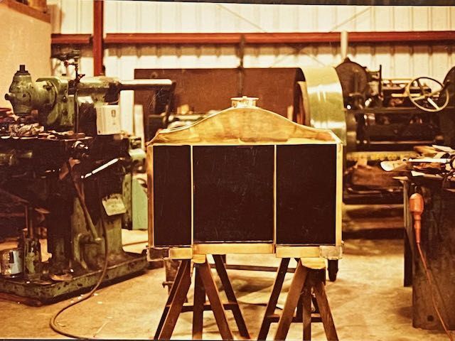 A brass-trimmed car radiator sits on wooden sawhorses in a cluttered workshop surrounded by industrial machinery.