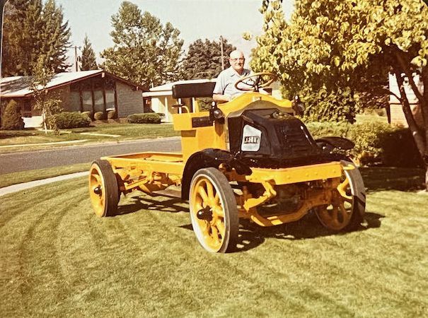 An elderly person sits at the wheel of a yellow, vintage truck chassis parked on a suburban lawn.