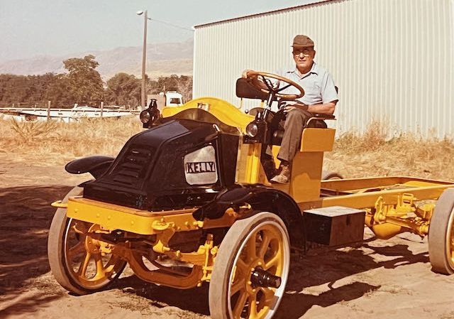 An individual sits on a yellow vintage truck chassis with a black radiator housing, parked in an outdoor, rural setting.