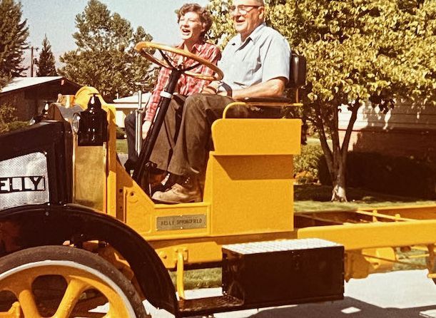An elderly couple sits laughing in a restored vintage yellow truck on a sunny day.