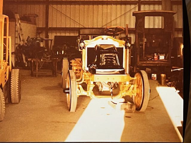 A vintage yellow car under restoration in a cluttered garage with a wooden cabin frame in the background.
