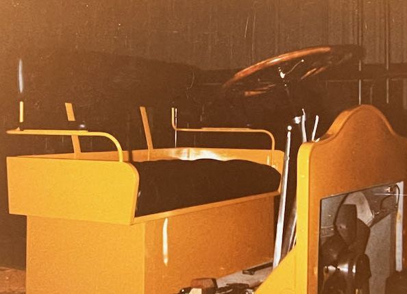 A vintage yellow, bench-seated golf cart or small vehicle with a wooden steering wheel, viewed from the side.