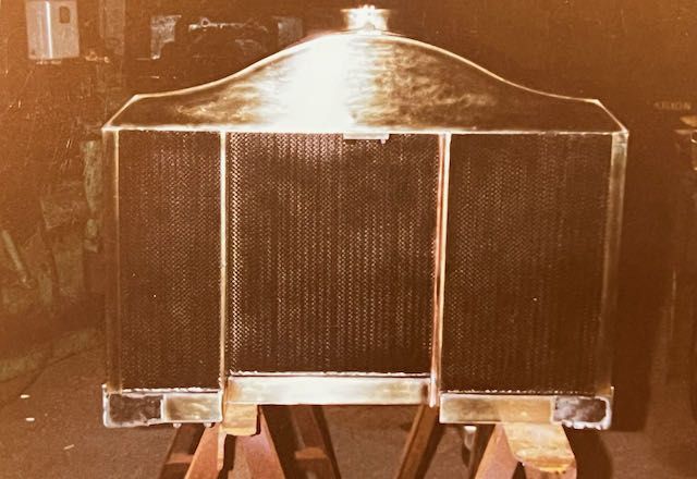 A vintage brass-toned radiator for an antique vehicle, resting on wooden sawhorses in a workshop setting.