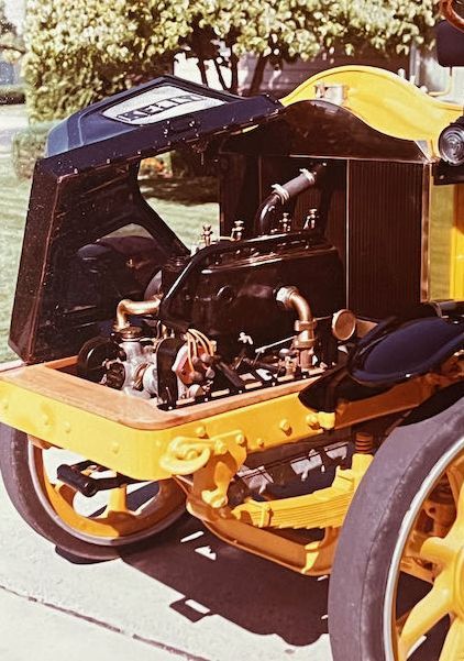 A bright yellow vintage automobile with its hood open, revealing the black engine block and brass plumbing.