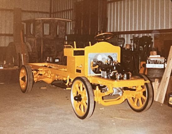 A bright yellow, partially restored vintage truck chassis with large spoked wheels, situated inside a cluttered workshop.