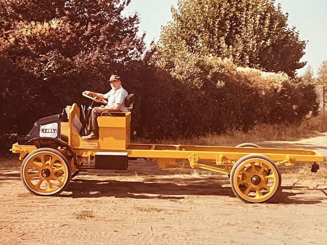A person sits in the driver’s seat of a bright yellow, vintage truck chassis with large spoke wheels outdoors.