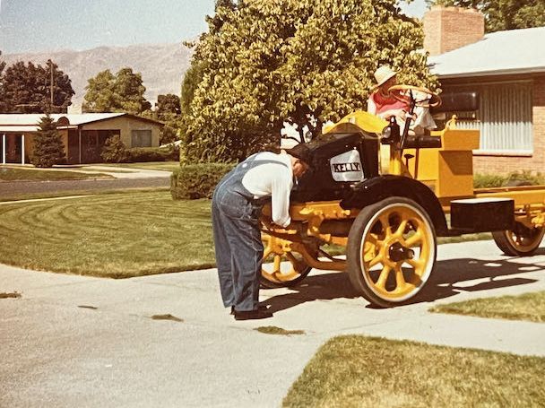 A person in overalls examines the engine of a vintage yellow vehicle parked on a residential driveway.