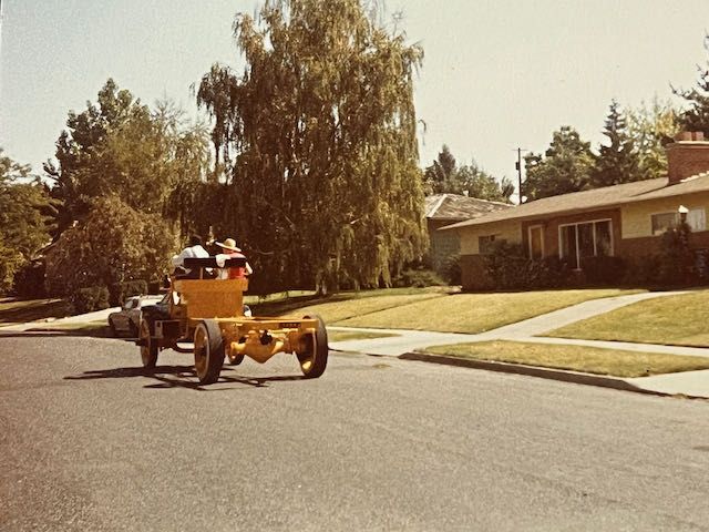 A vintage yellow open-frame vehicle carrying passengers drives down a residential street past a house and large trees.