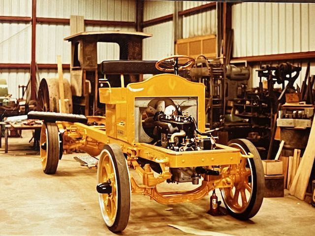 A bright yellow antique vehicle chassis with exposed engine and wooden wheels sits in a workshop.