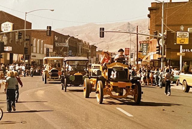 A vintage car parade proceeds down a sunlit city street, framed by brick buildings and mountains in the background.