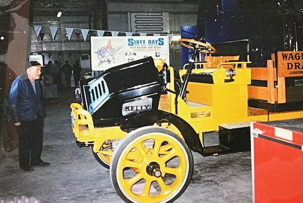 An older person looks at a yellow and black vintage Kelly truck parked inside an exhibit hall.