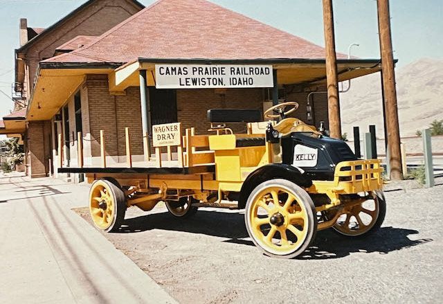 A bright yellow vintage flatbed truck displayed indoors, featuring spoked wheels and a wooden passenger-side crate.