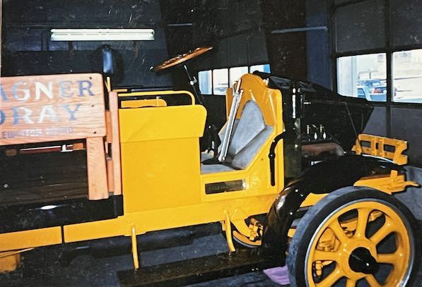 A bright yellow vintage truck displayed indoors, featuring large, spoked wheels and a wooden side panel.