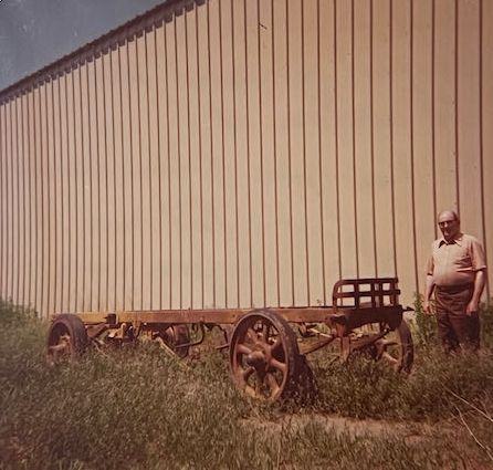 A person stands next to the rusted metal chassis and wheels of an antique wagon beside a tall, vertical-sided building.