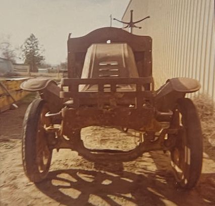 A rusted, vintage vehicle chassis missing its body, viewed from the front on a dirt lot beside a metal building.