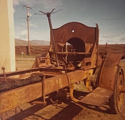 The rusted frame and front cowl of an antique vehicle sit outdoors on dirt ground under a blue sky.