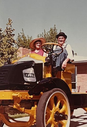 An antique black flatbed truck with large, bright red spoked wheels parked on a grassy field with other vintage trucks.