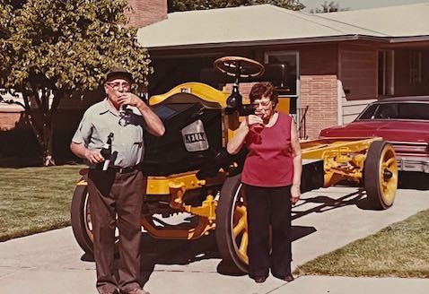 An older couple stands outdoors on a driveway in front of a restored yellow vintage vehicle, pouring a drink into a glass.