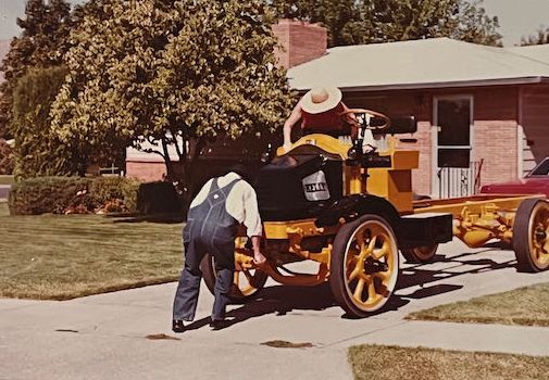 Two people work on a yellow, vintage truck chassis parked in a residential driveway.