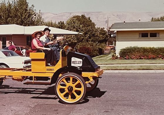 Two people in vintage hats sit on a restored yellow Kelly truck chassis on a sunny street next to suburban houses.