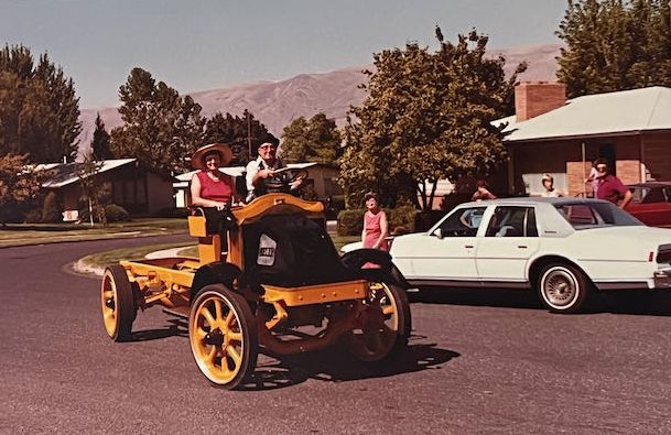 Two people ride a vintage, yellow, open-frame vehicle on a suburban street near a white sedan.