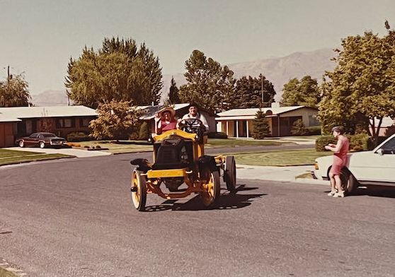 A yellow vintage flatbed truck with a driver and passenger drives past an old brick building during a sunny parade.