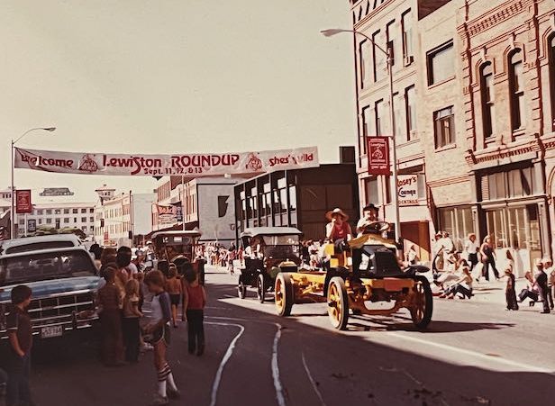 A vintage photograph shows a Lewiston Roundup parade on a city street with people and antique vehicles.