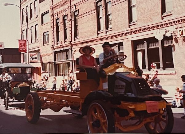 A yellow, vintage open-frame truck with two people driving through a parade in front of historic brick buildings.